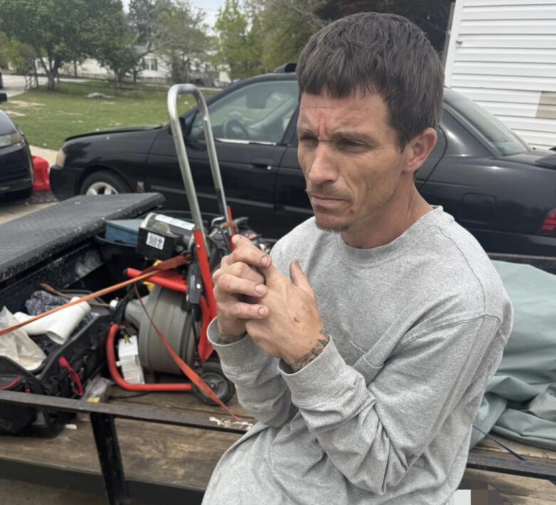 A man with short dark hair wearing a light gray long-sleeve shirt and tan cargo pants is sitting on the edge of a utility trailer. The trailer contains various tools and equipment, including a red dolly and a coiled hose. The man has a tattoo on his left wrist and appears to be clasping his hands together while looking to the side with a serious expression. A black car is parked behind the trailer, and there is a sheriff's badge logo in the bottom left corner of the image.