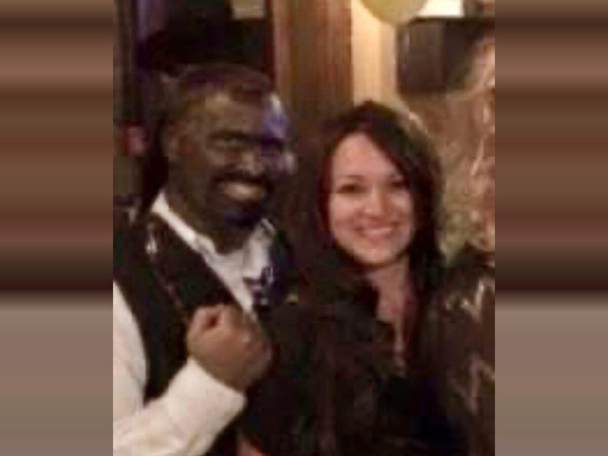 A man and a woman standing close together, both smiling. The man is wearing a white shirt with a dark vest, and the woman has long dark hair and is wearing a dark top. The background appears to be indoors with warm lighting.