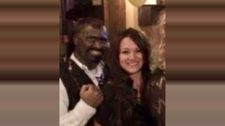 A man and a woman standing close together, both smiling. The man is wearing a white shirt with a dark vest, and the woman has long dark hair and is wearing a dark top. The background appears to be indoors with warm lighting.