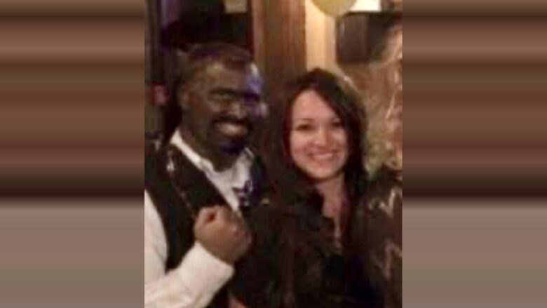 A man and a woman standing close together, both smiling. The man is wearing a white shirt with a dark vest, and the woman has long dark hair and is wearing a dark top. The background appears to be indoors with warm lighting.