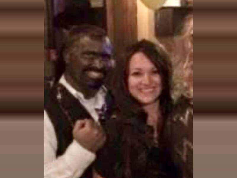 A man and a woman standing close together, both smiling. The man is wearing a white shirt with a dark vest, and the woman has long dark hair and is wearing a dark top. The background appears to be indoors with warm lighting.