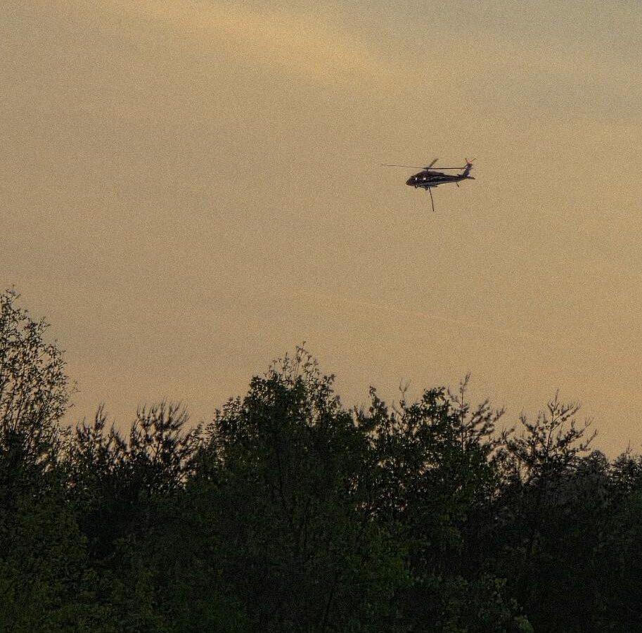 A helicopter flying above a dense forest with a sky that has a warm, muted tone, suggesting either early morning or late evening. The helicopter is dark-colored and appears to be hovering or moving slowly. The trees below are lush and green.