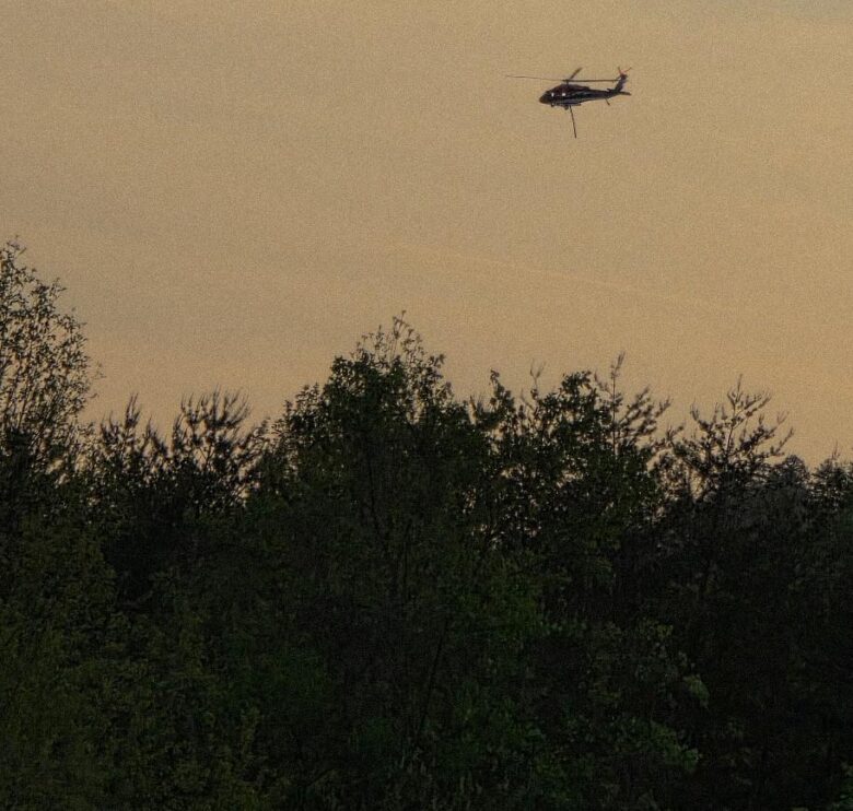 A helicopter flying above a dense forest with a sky that has a warm, muted tone, suggesting either early morning or late evening. The helicopter is dark-colored and appears to be hovering or moving slowly. The trees below are lush and green.