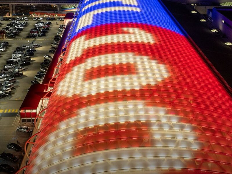 A large, curved building roof illuminated at night with red, white, and blue lights forming the words "ATL" and "ATLANTA" in a bold, stylized font. The surrounding area includes a parking lot filled with cars and other buildings in the background. The bottom left corner features the text "ATL Pic of the Week" in red and yellow.