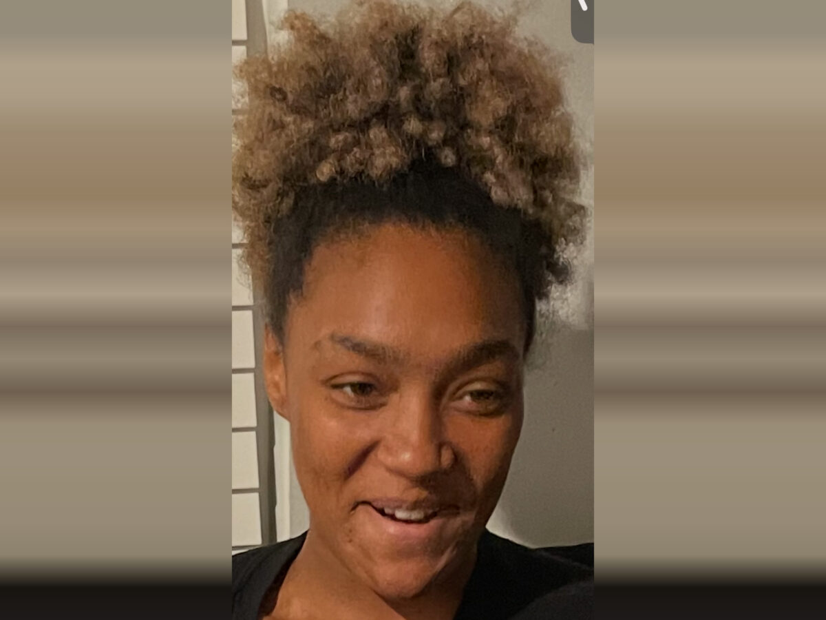 A close-up of a person with curly, light brown hair pulled up into a high puff. They have a medium skin tone and are wearing a black top. The person is smiling slightly and looking slightly to the side. The background is neutral with a hint of a window or blinds on the left side.