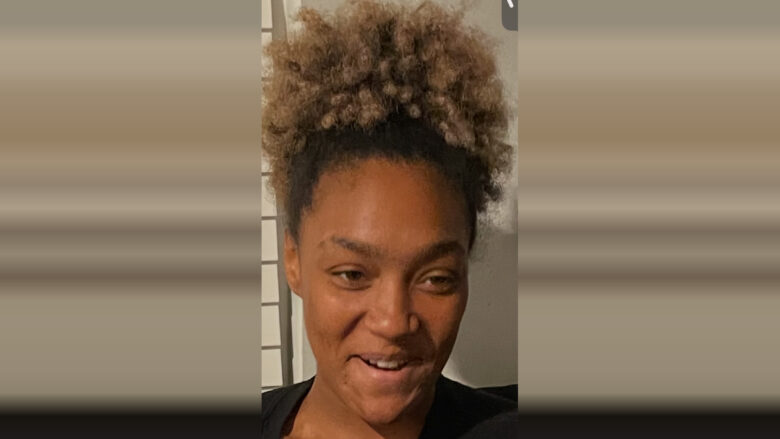 A close-up of a person with curly, light brown hair pulled up into a high puff. They have a medium skin tone and are wearing a black top. The person is smiling slightly and looking slightly to the side. The background is neutral with a hint of a window or blinds on the left side.