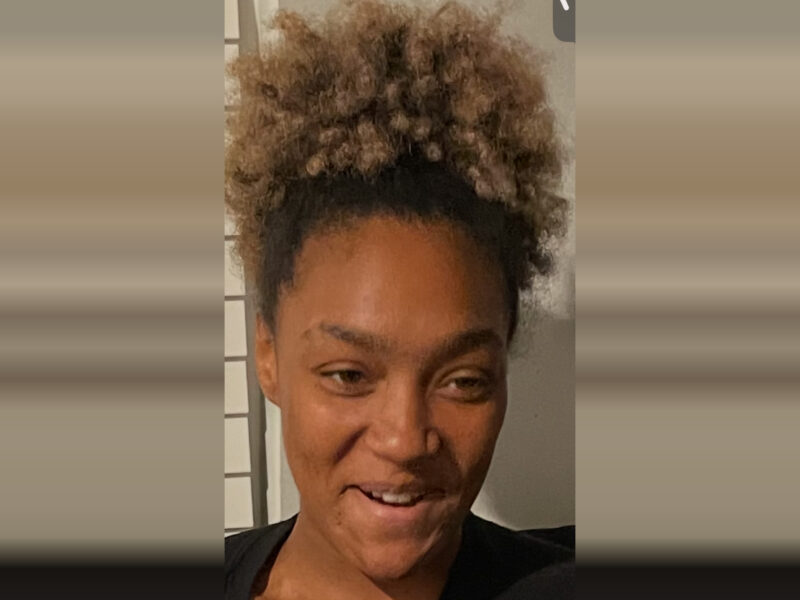 A close-up of a person with curly, light brown hair pulled up into a high puff. They have a medium skin tone and are wearing a black top. The person is smiling slightly and looking slightly to the side. The background is neutral with a hint of a window or blinds on the left side.