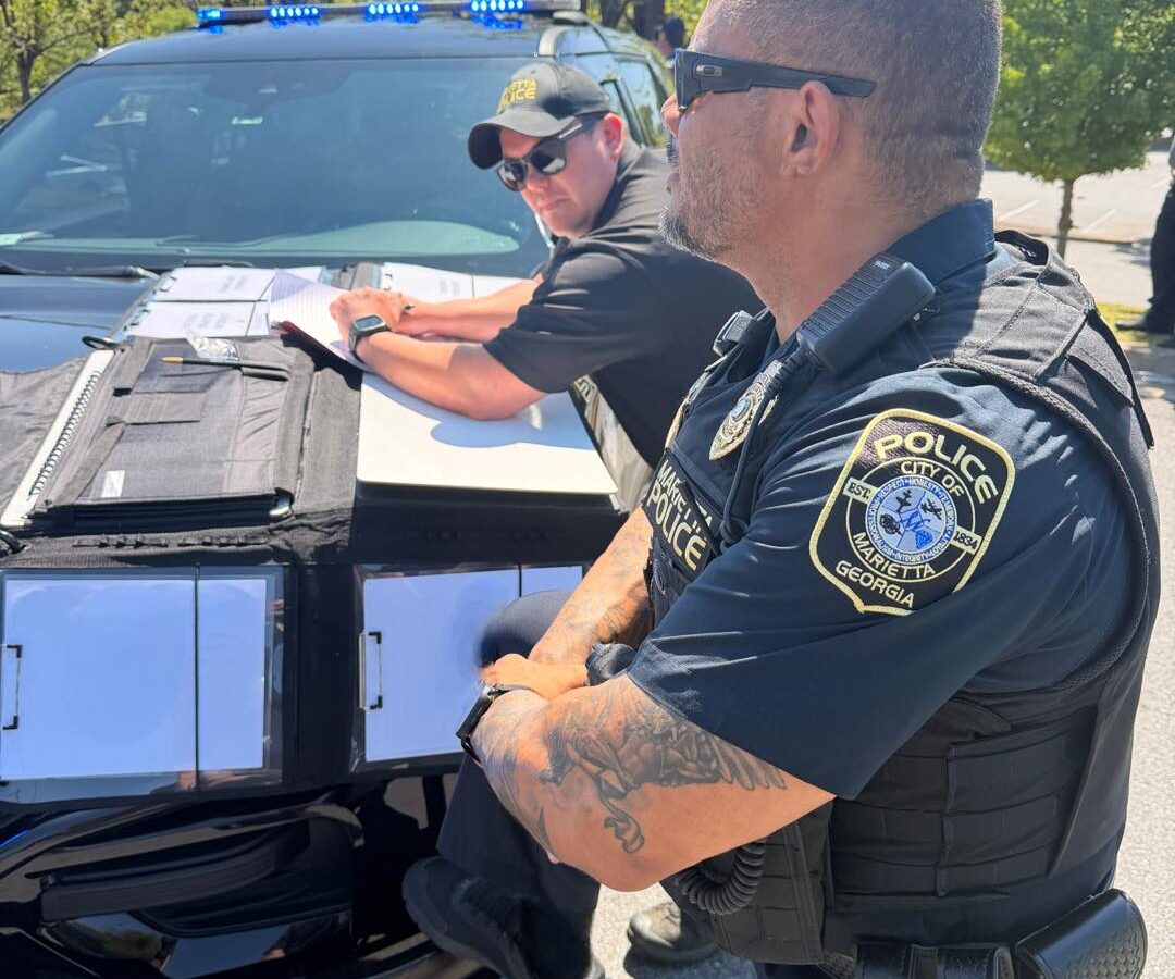 Two police officers from Marietta, Georgia, are engaged in work outdoors beside a black police vehicle with blue lights on top. One officer, wearing sunglasses and a cap, is leaning on the hood of the car, writing on papers placed on a folder. The other officer, also wearing sunglasses and a tactical vest, stands with arms crossed, showing a detailed tattoo on his forearm. Trees and a clear sky are visible in the background.