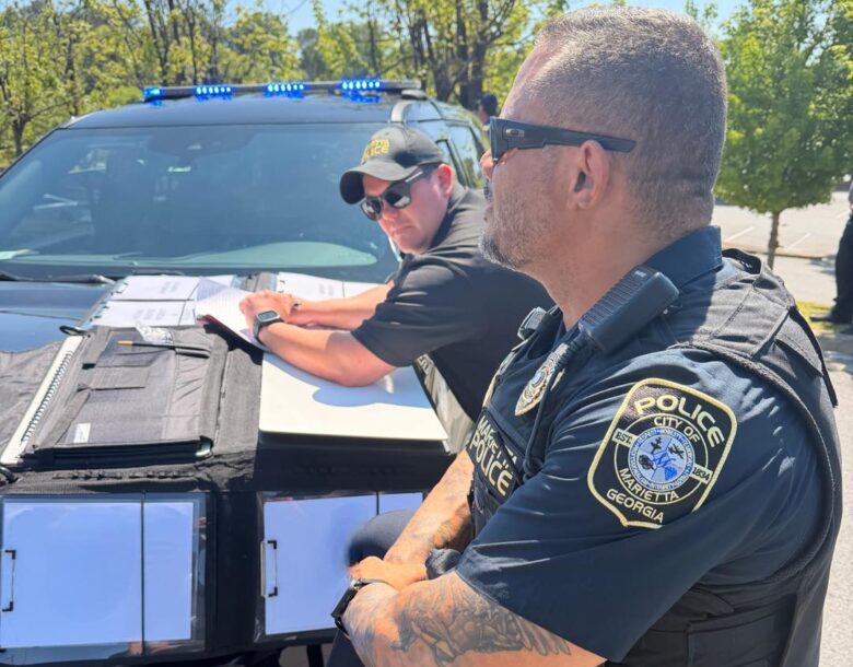 Two police officers from Marietta, Georgia, are engaged in work outdoors beside a black police vehicle with blue lights on top. One officer, wearing sunglasses and a cap, is leaning on the hood of the car, writing on papers placed on a folder. The other officer, also wearing sunglasses and a tactical vest, stands with arms crossed, showing a detailed tattoo on his forearm. Trees and a clear sky are visible in the background.