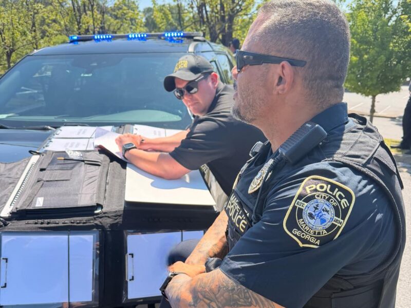 Two police officers from Marietta, Georgia, are engaged in work outdoors beside a black police vehicle with blue lights on top. One officer, wearing sunglasses and a cap, is leaning on the hood of the car, writing on papers placed on a folder. The other officer, also wearing sunglasses and a tactical vest, stands with arms crossed, showing a detailed tattoo on his forearm. Trees and a clear sky are visible in the background.