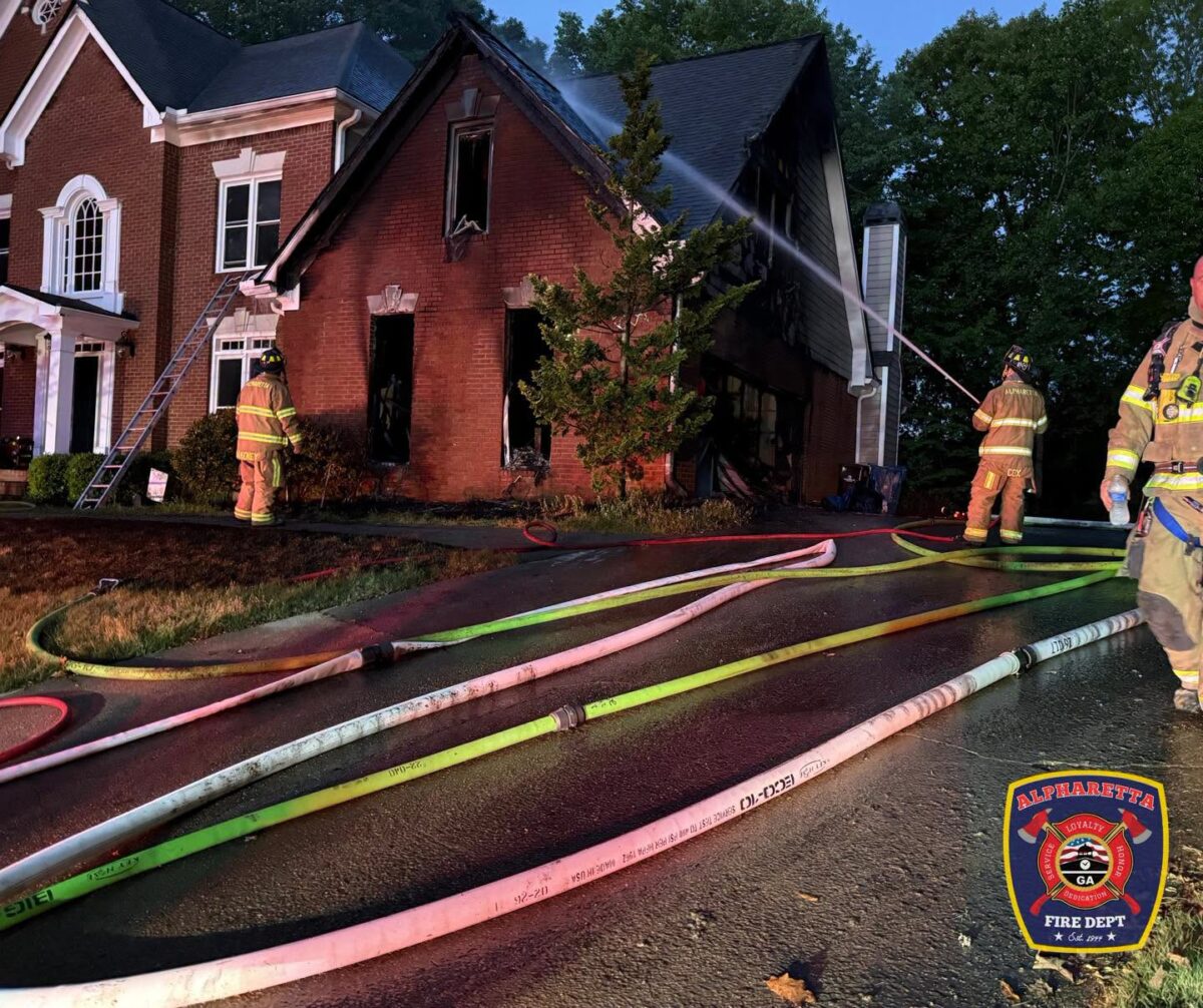 A brick house with significant fire damage, especially on the right side, is being sprayed with water by firefighters. Several fire hoses are laid out on the wet driveway in front of the house. Three firefighters in full gear are visible, with one actively spraying water on the house. The scene is set during dusk or early night, and the Alpharetta Fire Department emblem is displayed in the bottom right corner.