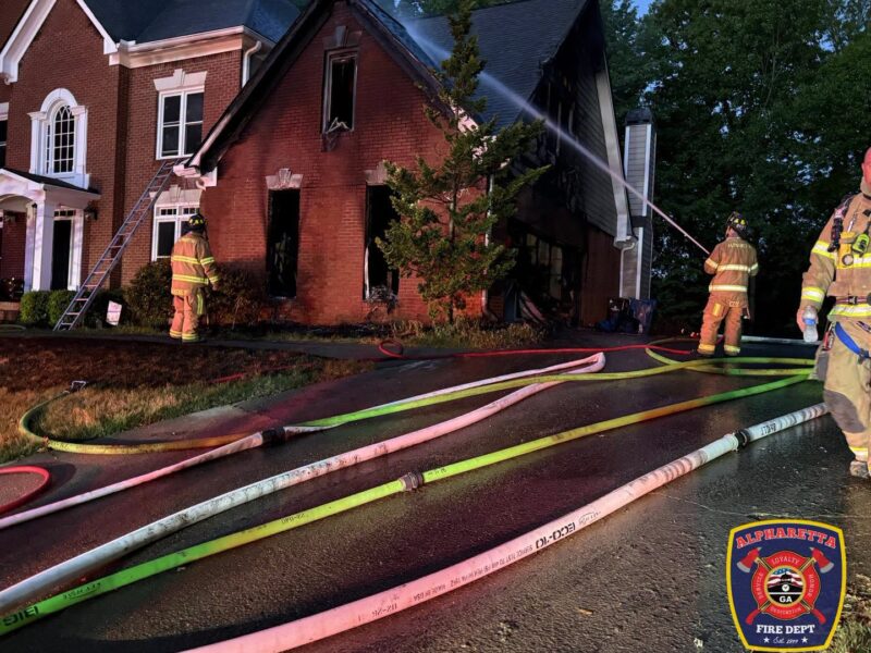 A brick house with significant fire damage, especially on the right side, is being sprayed with water by firefighters. Several fire hoses are laid out on the wet driveway in front of the house. Three firefighters in full gear are visible, with one actively spraying water on the house. The scene is set during dusk or early night, and the Alpharetta Fire Department emblem is displayed in the bottom right corner.
