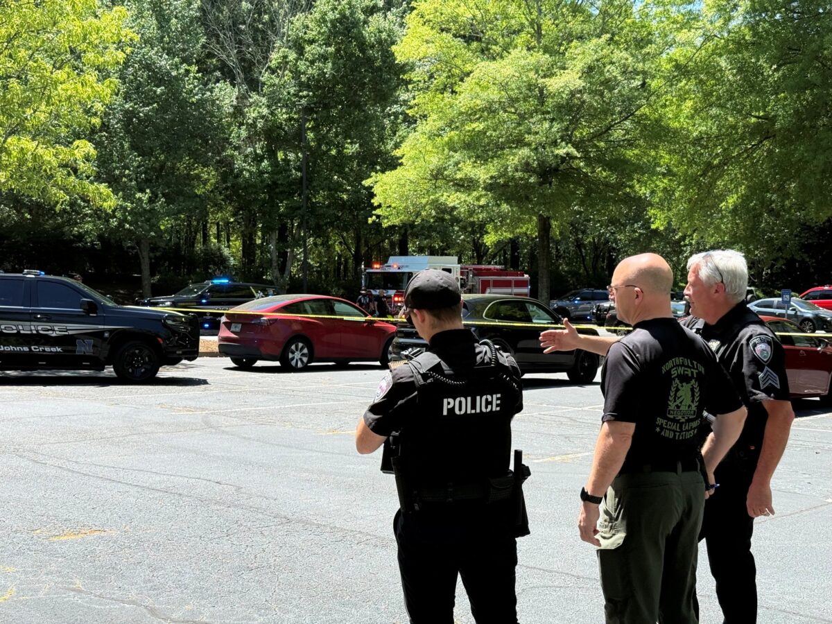Three law enforcement officers stand in a parking lot near yellow police tape. One officer wears a black vest labeled "POLICE," another wears a black shirt with "NORTHFULTON SWAT" on the back, and the third wears a black uniform with a sergeant patch. Several police vehicles with flashing lights and a fire truck are visible in the background, along with trees and parked cars.