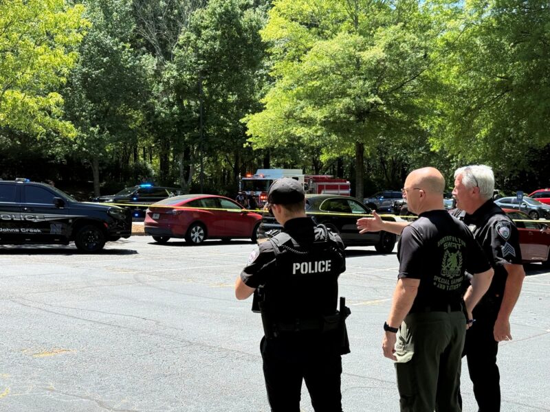 Three law enforcement officers stand in a parking lot near yellow police tape. One officer wears a black vest labeled "POLICE," another wears a black shirt with "NORTHFULTON SWAT" on the back, and the third wears a black uniform with a sergeant patch. Several police vehicles with flashing lights and a fire truck are visible in the background, along with trees and parked cars.