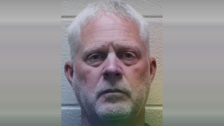 Middle-aged man with short white hair and light facial stubble, looking directly at the camera with a neutral expression, against a light-colored brick wall background.