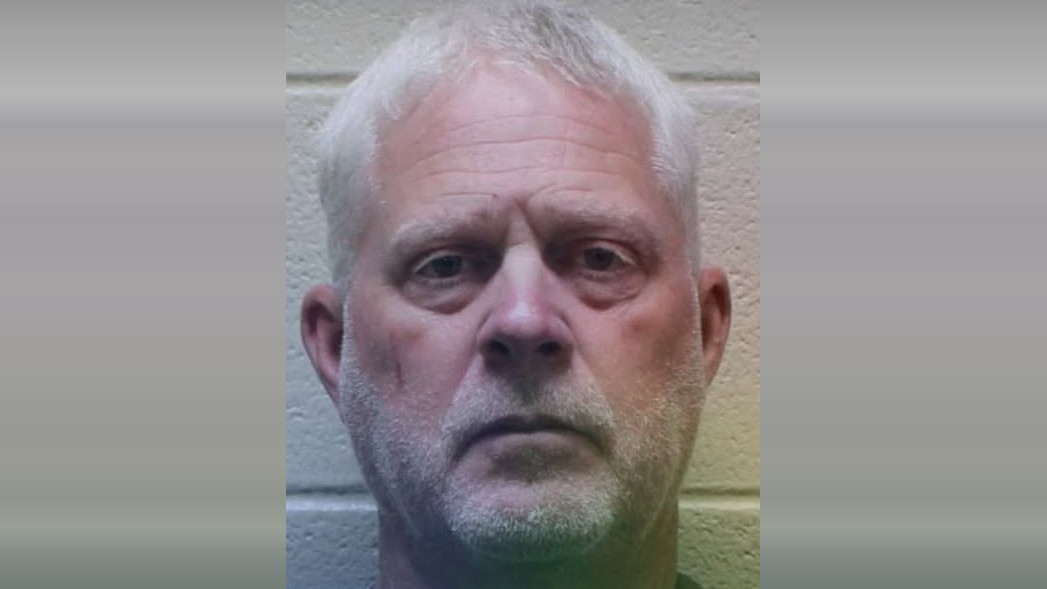 Middle-aged man with short white hair and light facial stubble, looking directly at the camera with a neutral expression, against a light-colored brick wall background.