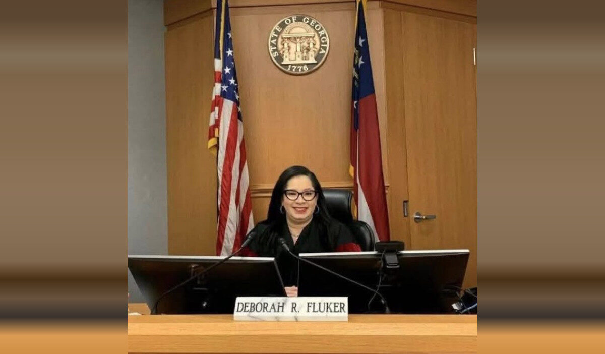 A woman with long dark hair and glasses is seated behind a judge's bench in a courtroom. She is wearing a black robe and smiling. In front of her is a nameplate that reads "Deborah R. Fluker." Behind her are two flags, one of the United States and one of Georgia, with the seal of the State of Georgia mounted on the wooden wall.
