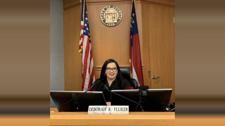 A woman with long dark hair and glasses is seated behind a judge's bench in a courtroom. She is wearing a black robe and smiling. In front of her is a nameplate that reads "Deborah R. Fluker." Behind her are two flags, one of the United States and one of Georgia, with the seal of the State of Georgia mounted on the wooden wall.