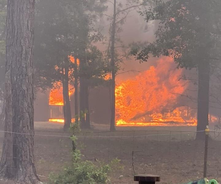 A large fire engulfs a building in a wooded area, with bright orange flames and thick smoke visible through the trees. In the foreground, firefighting equipment, including hoses and a valve, is positioned on the ground.