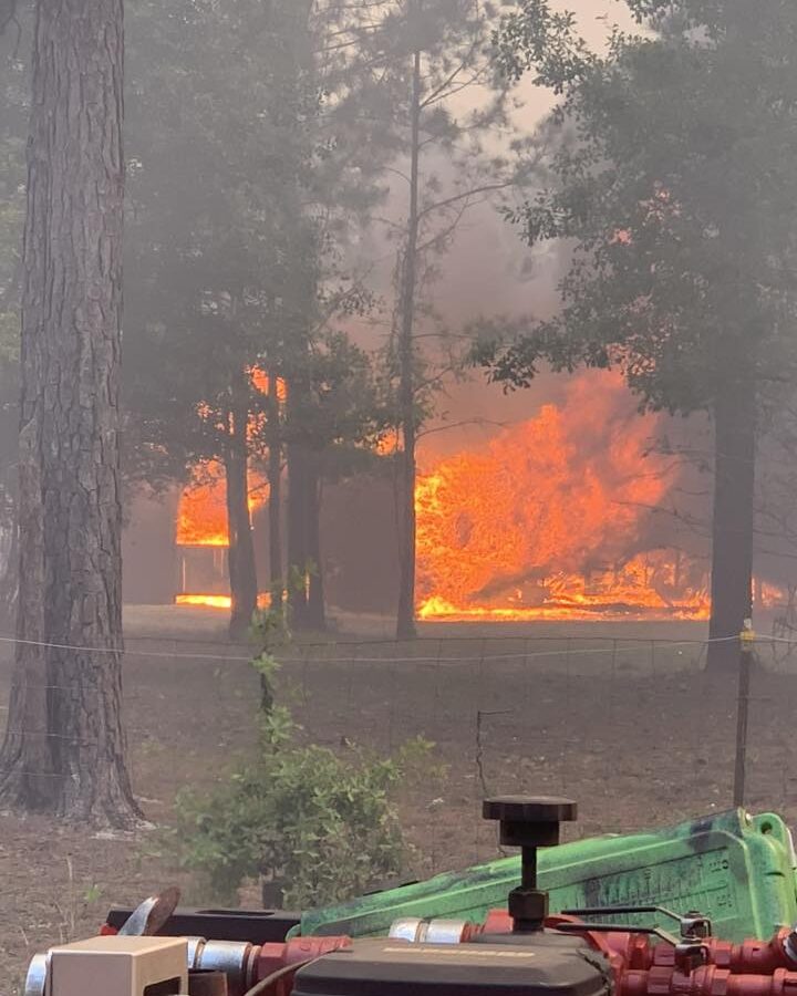 A large fire engulfs a building in a wooded area, with bright orange flames and thick smoke visible through the trees. In the foreground, firefighting equipment, including hoses and a valve, is positioned on the ground.