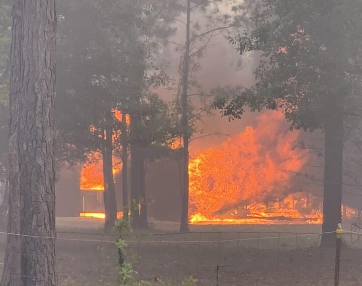A large fire engulfs a building in a wooded area, with bright orange flames and thick smoke visible through the trees. In the foreground, firefighting equipment, including hoses and a valve, is positioned on the ground.