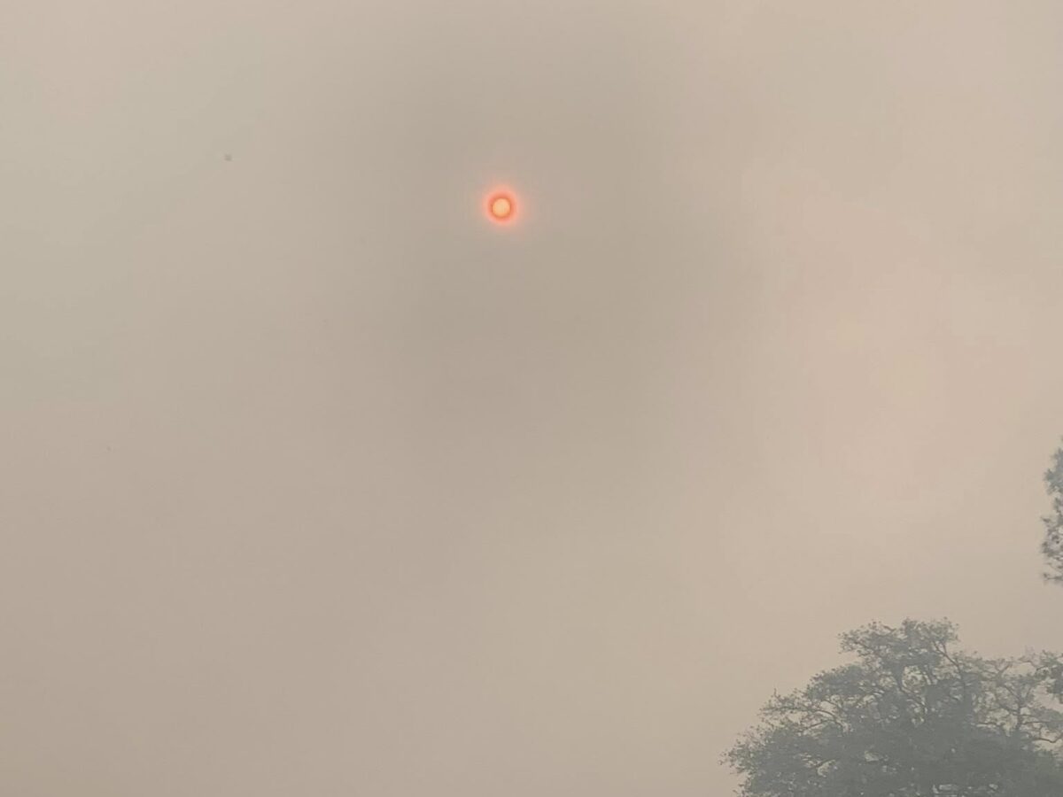 A construction backhoe loader parked in front of a small building with a metal roof. The sky is filled with thick smoke, giving it a hazy, orange appearance, and the sun appears as a small, bright red circle. There are trees partially visible through the smoke in the background.