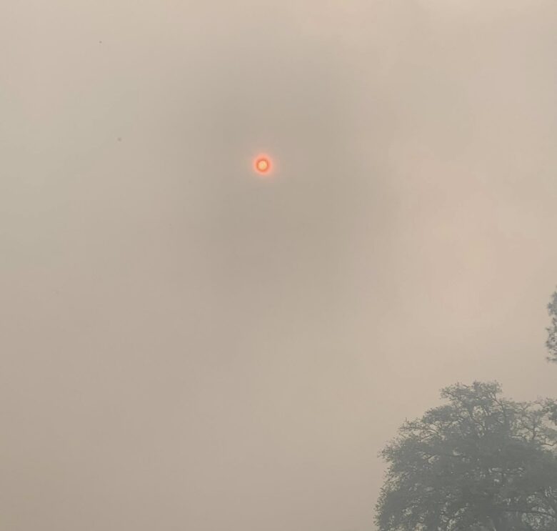 A construction backhoe loader parked in front of a small building with a metal roof. The sky is filled with thick smoke, giving it a hazy, orange appearance, and the sun appears as a small, bright red circle. There are trees partially visible through the smoke in the background.