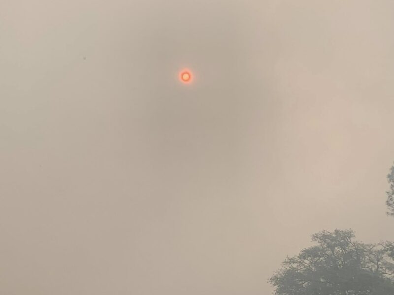A construction backhoe loader parked in front of a small building with a metal roof. The sky is filled with thick smoke, giving it a hazy, orange appearance, and the sun appears as a small, bright red circle. There are trees partially visible through the smoke in the background.