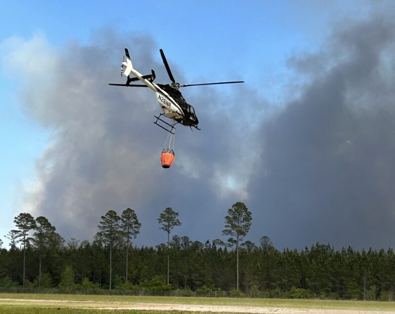 Helicopter carrying an orange water bucket flying over a grassy field with a forest and thick smoke in the background.