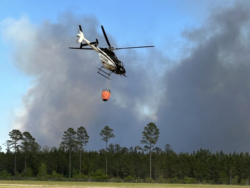 Helicopter carrying an orange water bucket flying over a grassy field with a forest and thick smoke in the background.