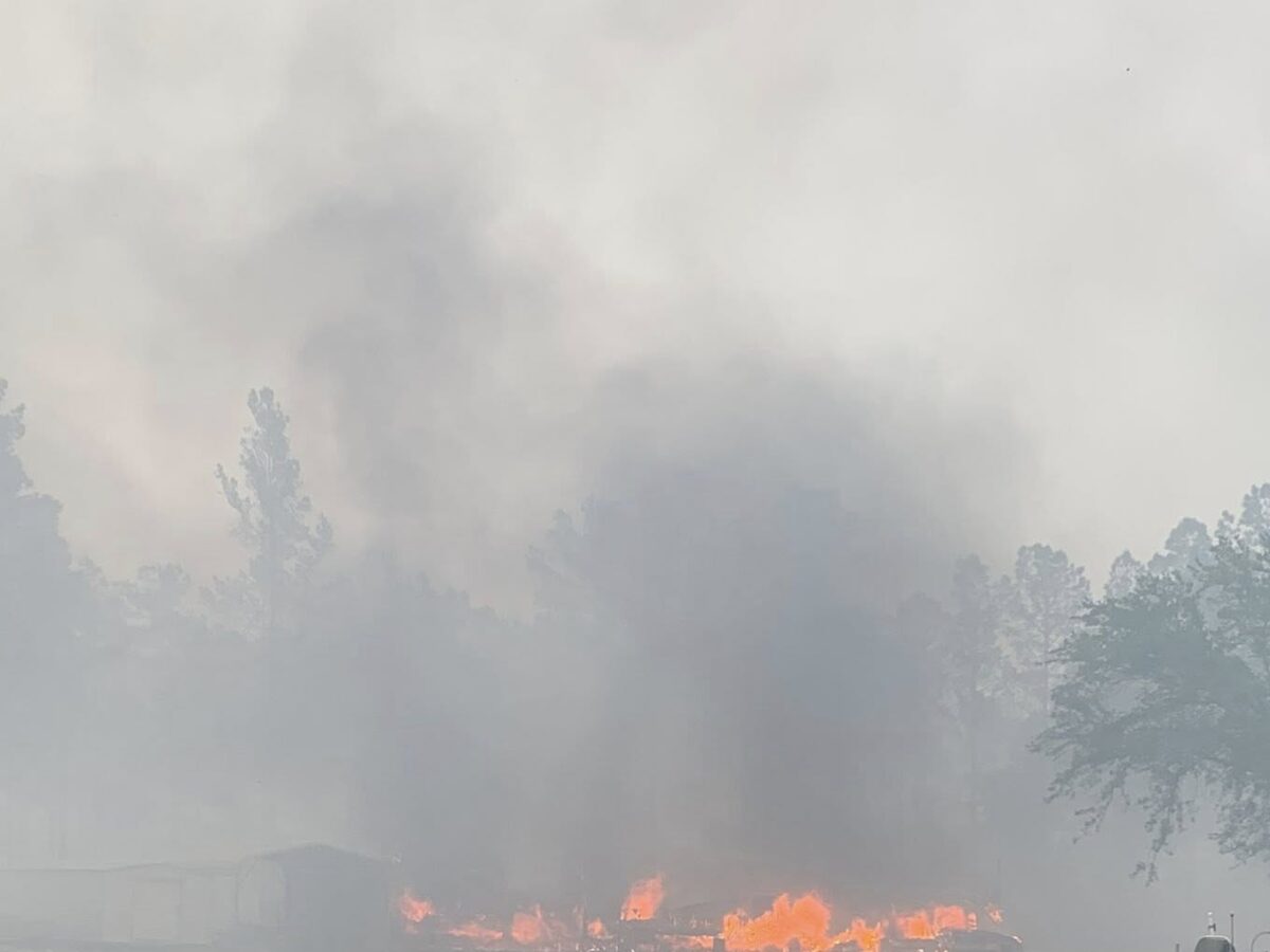 A fire burning intensely in the distance with thick smoke rising, partially obscuring trees and structures. The foreground shows scorched ground and patches of grass, with a small boat on a trailer positioned to the right.