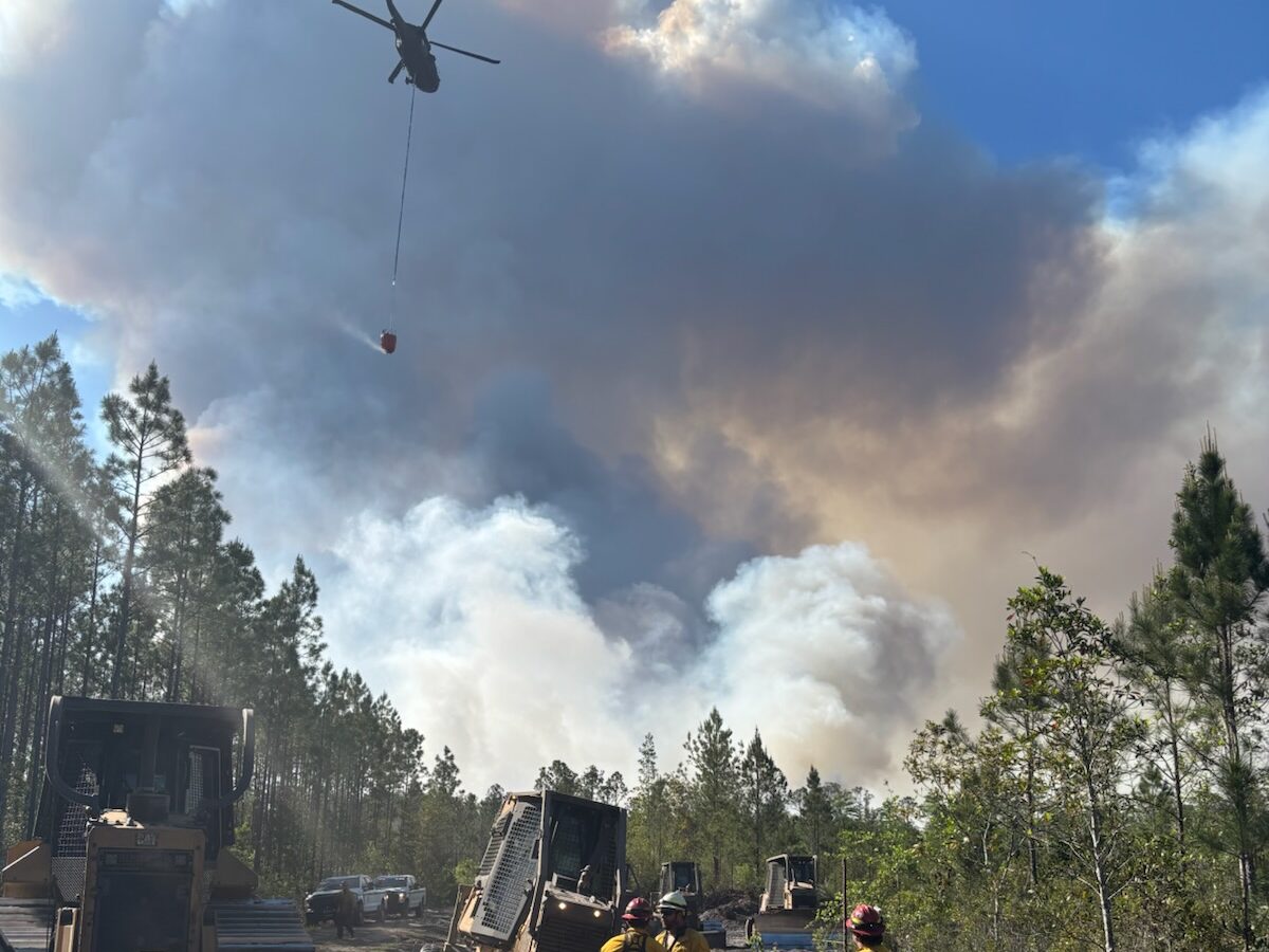Firefighters in yellow protective gear stand near bulldozers on a dirt path in a forested area. A helicopter carrying a water bucket flies above, releasing water onto a large plume of smoke rising from a wildfire in the background. The sky is partly clear with blue visible beyond the smoke.