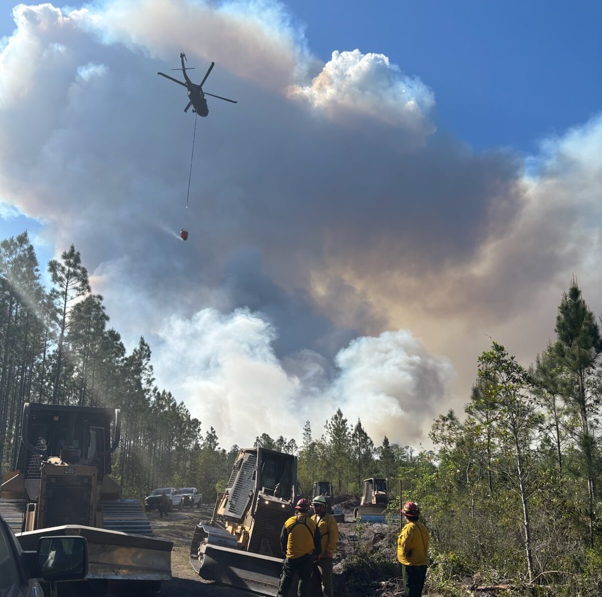 Firefighters in yellow protective gear stand near bulldozers on a dirt path in a forested area. A helicopter carrying a water bucket flies above, releasing water onto a large plume of smoke rising from a wildfire in the background. The sky is partly clear with blue visible beyond the smoke.