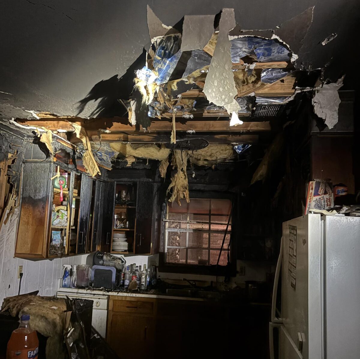 A severely fire-damaged kitchen with a large hole in the ceiling exposing insulation and wooden beams. The cabinets and walls are charred and blackened. A white refrigerator stands on the right side with some items on top, and a table with a large orange Fanta bottle is visible in the foreground. The window at the back is partially obscured by soot and damage.