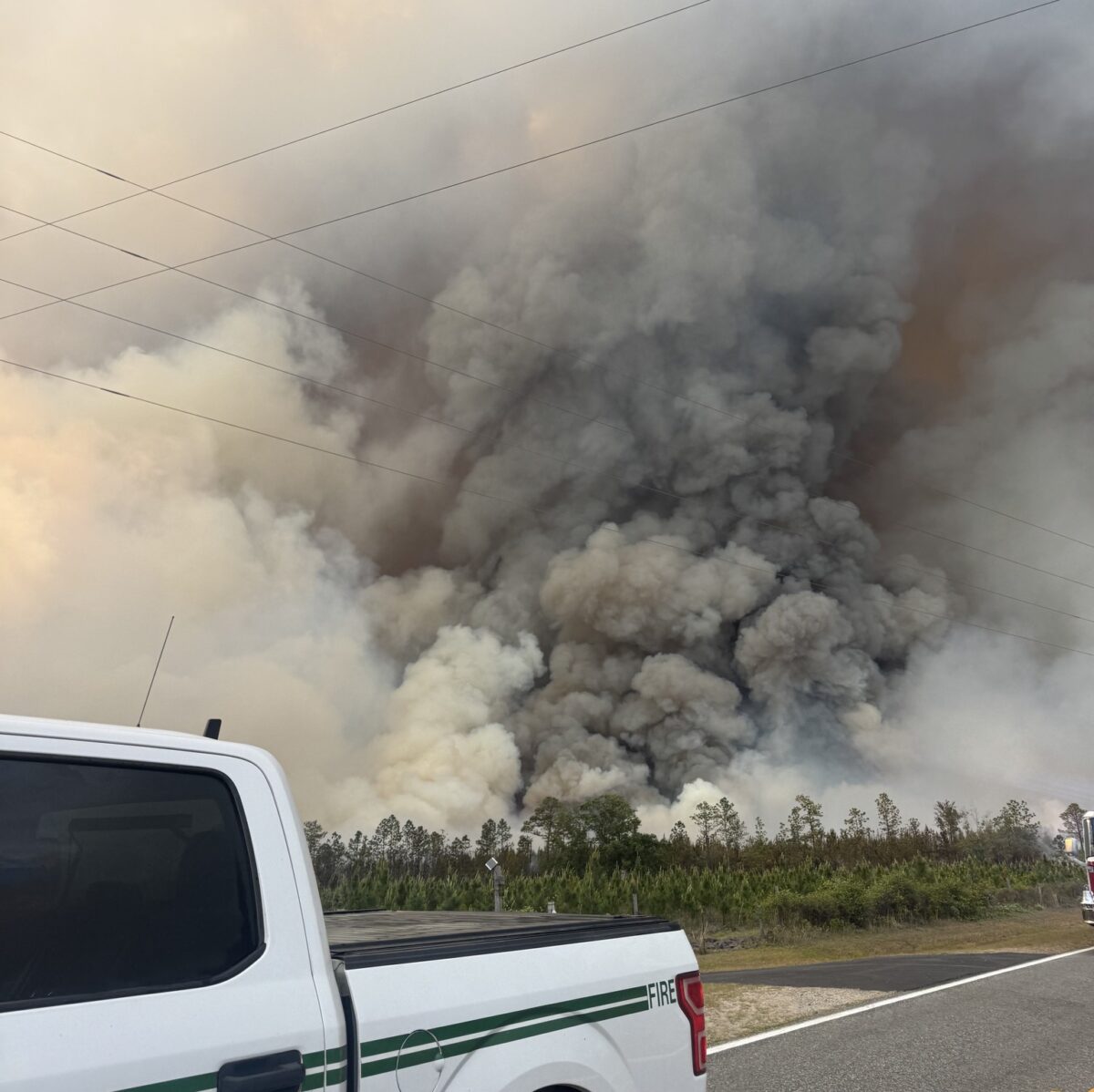 A white pickup truck with green stripes and the word "FIRE" partially visible on the side is parked on a road. In the background, there is a large, dense cloud of smoke rising from a fire in a wooded area, with trees visible at the base of the smoke. Power lines stretch across the sky above the smoke.