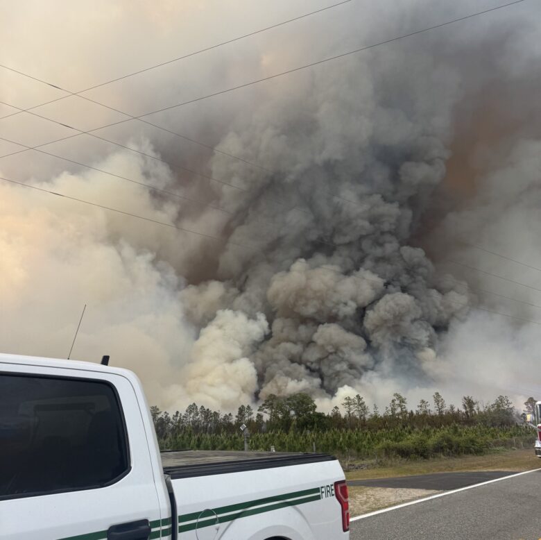 A white pickup truck with green stripes and the word "FIRE" partially visible on the side is parked on a road. In the background, there is a large, dense cloud of smoke rising from a fire in a wooded area, with trees visible at the base of the smoke. Power lines stretch across the sky above the smoke.