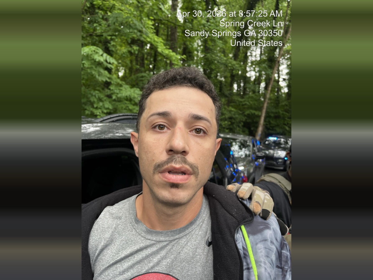 A close-up of a man with short curly hair, a mustache, and a small goatee, wearing a gray t-shirt and a black jacket with a gray and green patterned sleeve. He appears to be outdoors in a wooded area with green trees in the background. A gloved hand is resting on his shoulder. Behind him, there are vehicles with flashing blue lights, suggesting a law enforcement presence. The image includes a timestamp and location text at the top right corner: "Apr 30, 2026 at 8:57:25 AM, Spring Creek Ln, Sandy Springs GA 30350, United States.