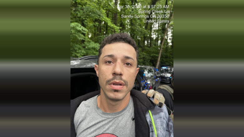 A close-up of a man with short curly hair, a mustache, and a small goatee, wearing a gray t-shirt and a black jacket with a gray and green patterned sleeve. He appears to be outdoors in a wooded area with green trees in the background. A gloved hand is resting on his shoulder. Behind him, there are vehicles with flashing blue lights, suggesting a law enforcement presence. The image includes a timestamp and location text at the top right corner: "Apr 30, 2026 at 8:57:25 AM, Spring Creek Ln, Sandy Springs GA 30350, United States.