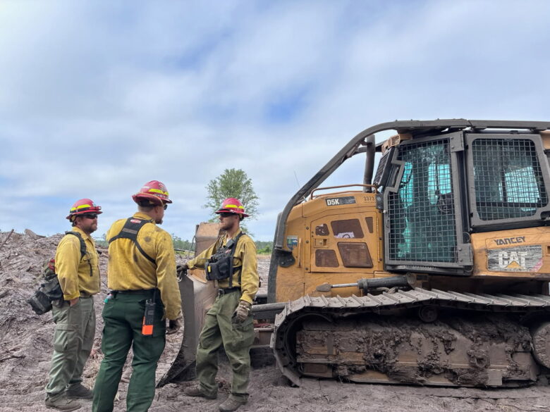Three workers wearing yellow shirts, green pants, red helmets, and safety gear stand next to a large yellow Caterpillar D5K bulldozer on muddy ground under a cloudy sky.