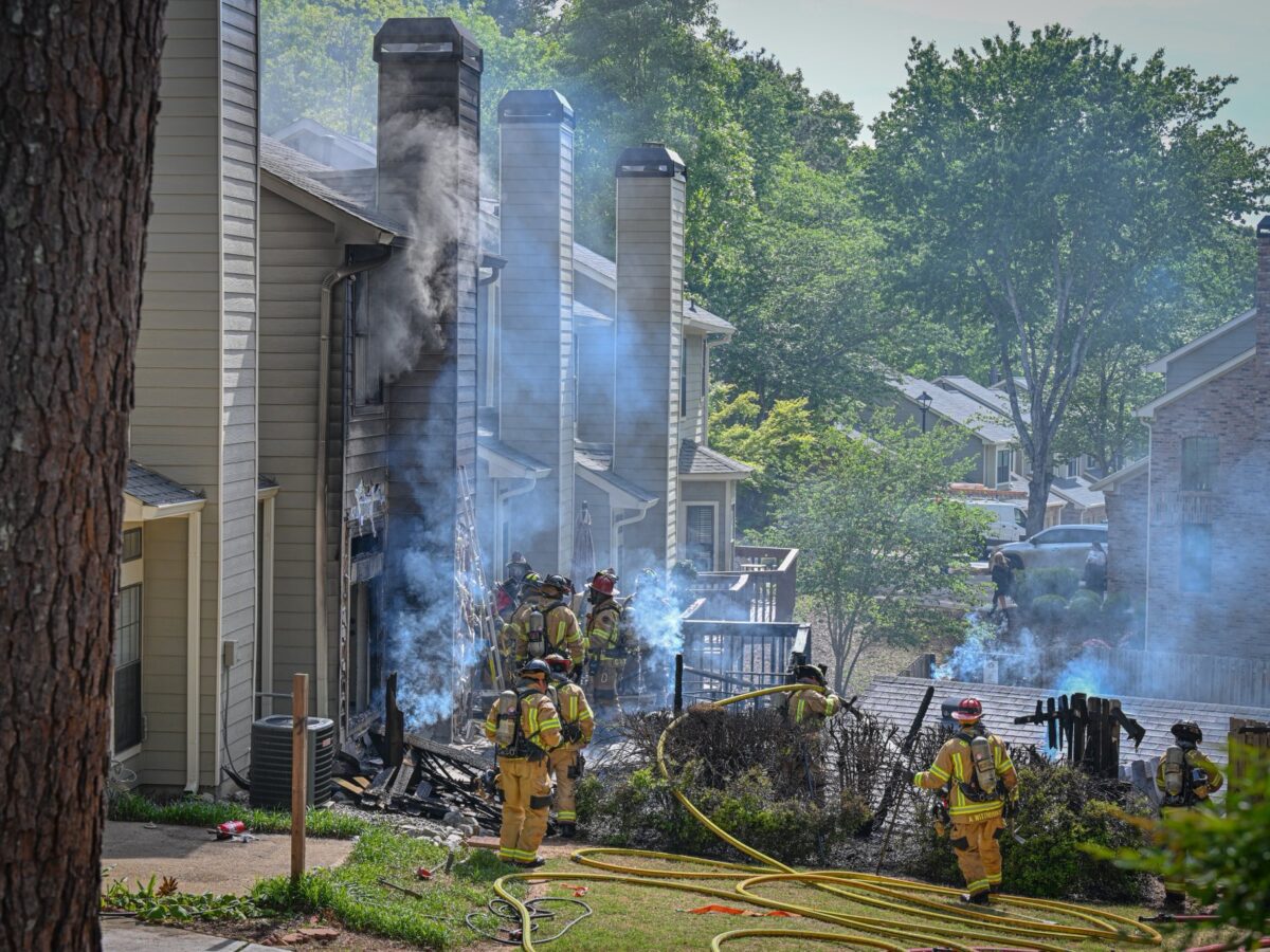 Firefighters in yellow protective gear are actively responding to a house fire in a residential area. Smoke is billowing from the side of a beige house with multiple chimneys, and the exterior shows signs of fire damage. Several fire hoses are laid out on the ground, and the firefighters are working near burnt bushes and debris. The scene is surrounded by green trees and neighboring houses.
