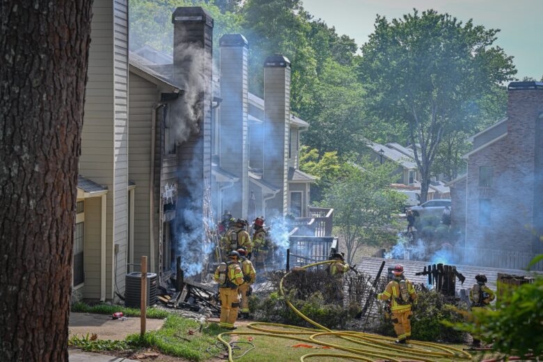 Firefighters in yellow protective gear are actively responding to a house fire in a residential area. Smoke is billowing from the side of a beige house with multiple chimneys, and the exterior shows signs of fire damage. Several fire hoses are laid out on the ground, and the firefighters are working near burnt bushes and debris. The scene is surrounded by green trees and neighboring houses.