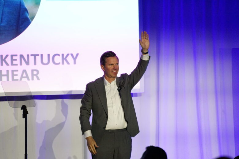 A man in a gray suit and white shirt is standing on a stage with a microphone clipped to his jacket, raising his right hand. Behind him is a screen with partially visible text that reads "KENTUCKY" and "HEAR." The background features blue and purple lighting with curtains.