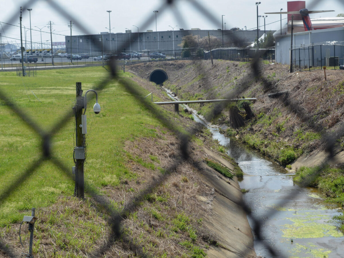 A narrow concrete drainage canal with some water and patches of green algae runs through a grassy and partially dry area. A wooden utility pole with a security camera is positioned near the canal. The scene is viewed through a chain-link fence, which is out of focus in the foreground. In the background, there are industrial buildings, parked vehicles, and a partial view of an airplane tail.