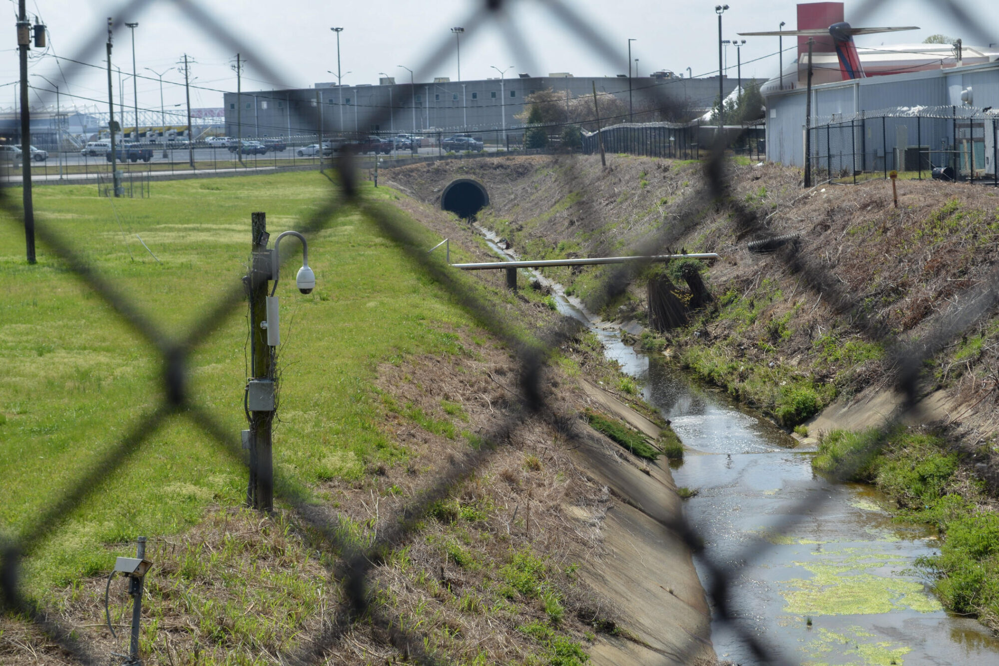A narrow concrete drainage canal with some water and patches of green algae runs through a grassy and partially dry area. A wooden utility pole with a security camera is positioned near the canal. The scene is viewed through a chain-link fence, which is out of focus in the foreground. In the background, there are industrial buildings, parked vehicles, and a partial view of an airplane tail.