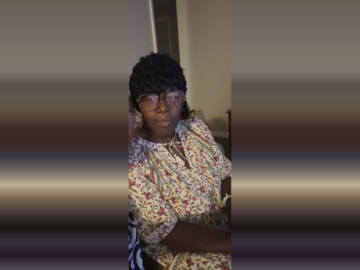 A person wearing large, square-framed glasses and a black textured hat. They are dressed in a light-colored blouse with a floral pattern featuring red and green accents. The background shows a beige wall and a partially open door.