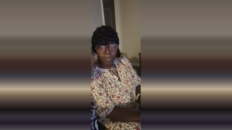 A person wearing large, square-framed glasses and a black textured hat. They are dressed in a light-colored blouse with a floral pattern featuring red and green accents. The background shows a beige wall and a partially open door.