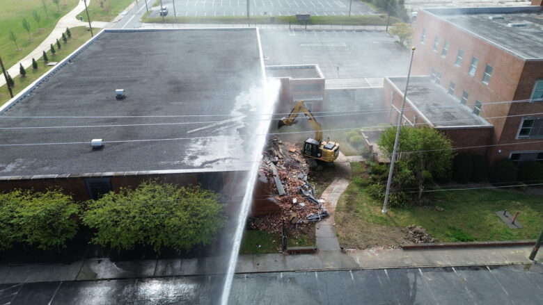 A yellow excavator is demolishing a brick building, creating a pile of rubble. Water is being sprayed onto the demolition site, likely to control dust. The building has a flat, dark roof and is surrounded by greenery and a sidewalk. There are power lines running across the image and an empty parking lot in the background.