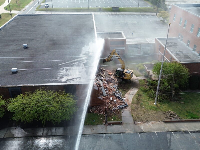 A yellow excavator is demolishing a brick building, creating a pile of rubble. Water is being sprayed onto the demolition site, likely to control dust. The building has a flat, dark roof and is surrounded by greenery and a sidewalk. There are power lines running across the image and an empty parking lot in the background.