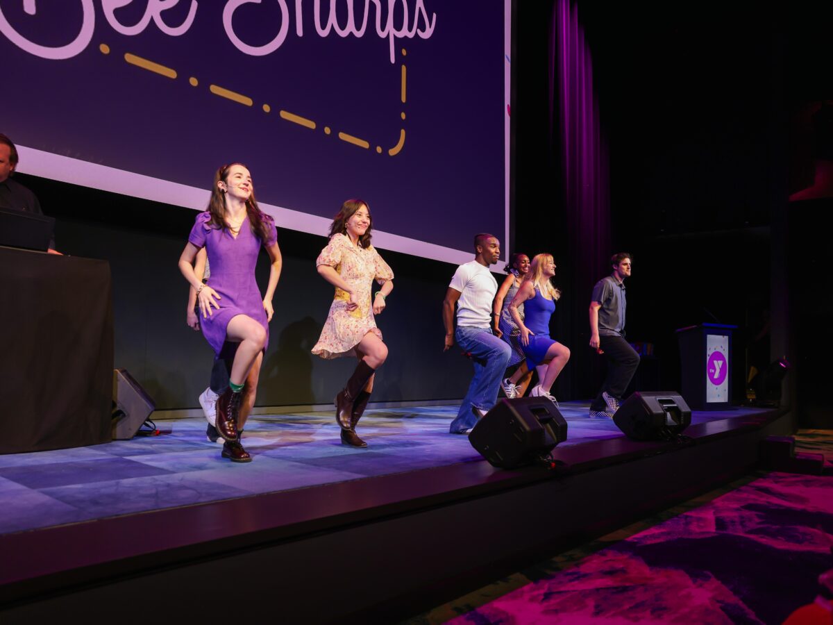 Six performers dancing on a stage under purple lighting, with a large screen behind them displaying partial text. The performers are mid-step, wearing casual and semi-formal attire, including dresses, boots, jeans, and sneakers. A man is visible at a DJ booth on the left side of the stage.