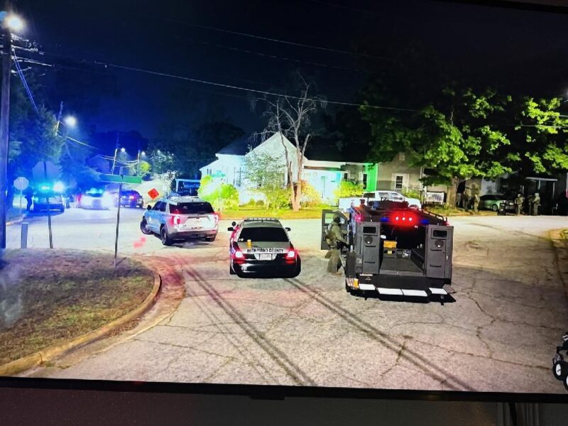 A nighttime scene on a residential street with multiple police vehicles, including a marked Richmond County police car and a large armored vehicle with its rear doors open. Several officers, some in tactical gear, are present near the vehicles and on the sidewalk. The area is illuminated by streetlights and the flashing blue and red lights of the police cars. Houses and trees line the street in the background.